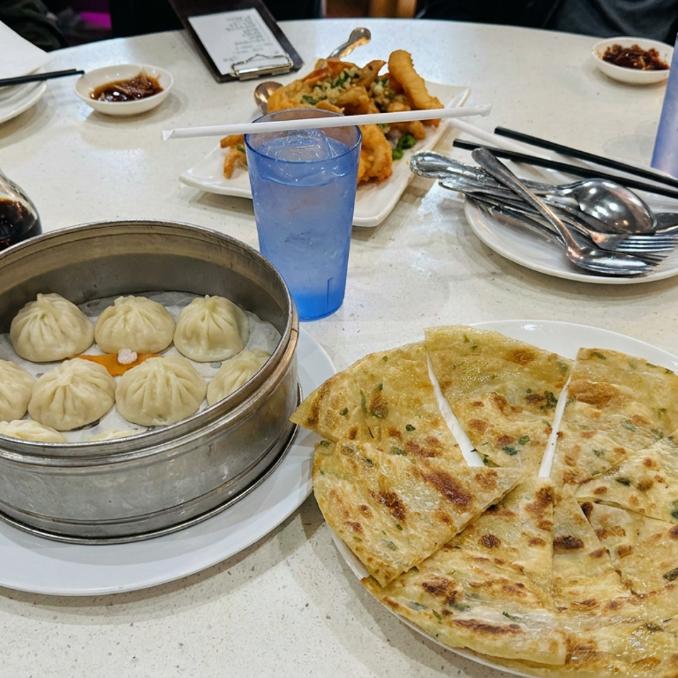 Chinese meal with steamed soup dumplings, sliced scallion pancake, fried appetizers, dipping sauces, chopsticks, and blue cups on a restaurant table.